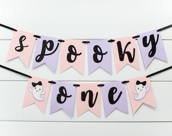 May include: A pink and purple banner with black glitter letters that reads "spooky one". The banner has two ghost cutouts with black bows.