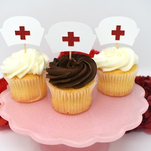 May include: Three cupcakes decorated with white and chocolate frosting, topped with red and white nurse hat cupcake toppers. The cupcakes are on a pink cake stand.
