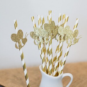 May include: Gold and white striped paper straws with gold glitter bee cutouts. The straws are in a white pitcher.