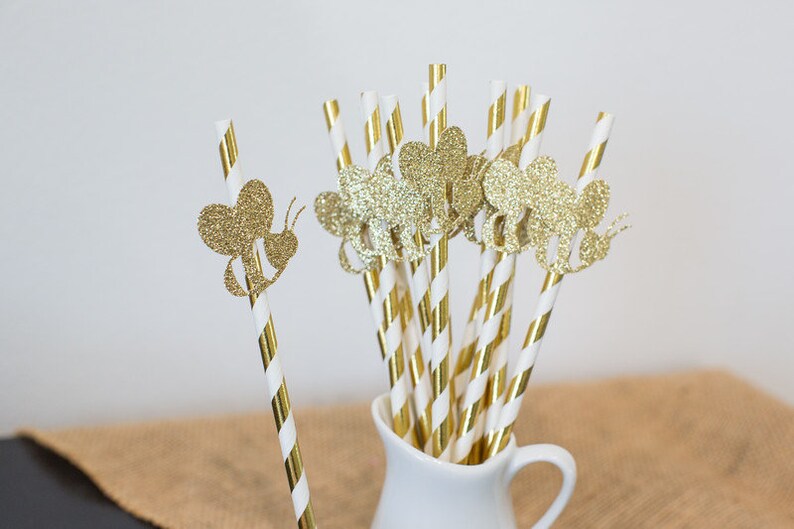 May include: Gold and white striped paper straws with gold glitter bee cutouts. The straws are in a white pitcher.