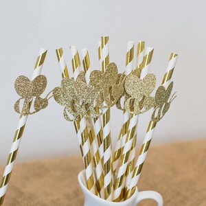 May include: Gold glitter straws with gold glitter heart cutouts. The straws are in a white pitcher.