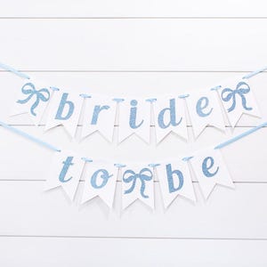 May include: A blue and white banner with the text "bride to be" in a light blue glitter font. The banner is strung with blue ribbon and has blue bows between each letter.