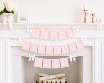 May include: A pink and white banner spelling out HAPPY BIRTHDAY ELLE, featuring rectangular pennants with white lettering on a light pink background, accented with small white bows on a pink ribbon string, displayed across a white fireplace mantel.