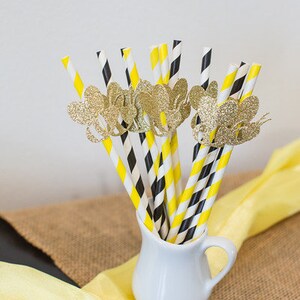 May include: A collection of decorative paper straws in a white pitcher. The straws have yellow and black stripes, with gold glitter butterfly accents. The pitcher sits on a burlap and yellow fabric background.