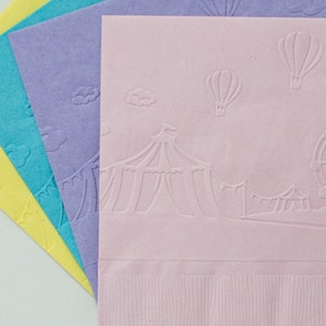 May include: Four pastel-colored paper napkins with an embossed design of a circus tent, hot air balloons, and a Ferris wheel. The napkins are yellow, light blue, lavender, and pink.