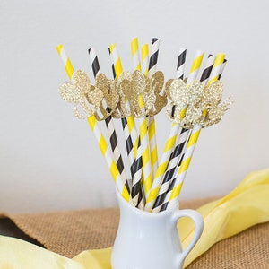 May include: A collection of decorative paper straws in a white pitcher. The straws have yellow and black stripes, and black and white diagonal stripes. Gold glitter butterfly embellishments adorn some of the straws. A yellow cloth and burlap are in the background.