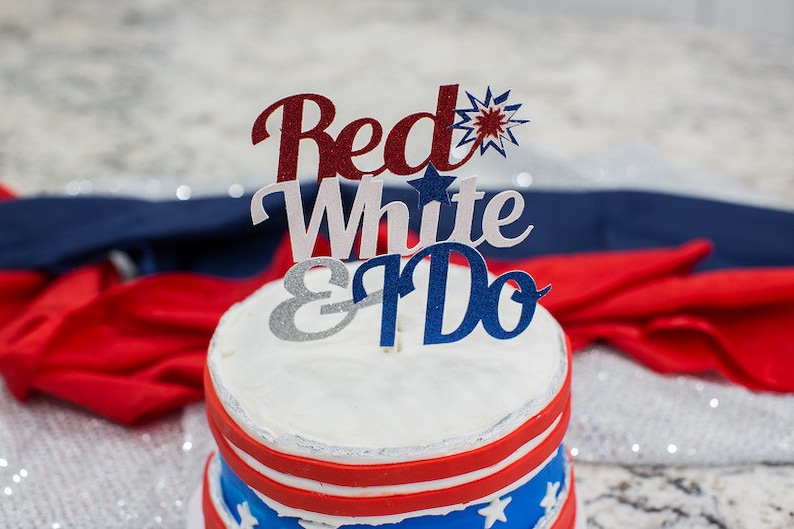 Pode incluir: Um bolo de casamento branco com cobertura vermelha, branca e azul e um topo de bolo que diz "Red White & Do".