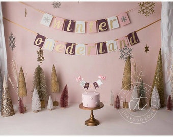 May include: A pink and gold winter wonderland themed cake with a banner that reads "winter wonderland one". The cake is decorated with snowflakes and is surrounded by small, sparkly trees.