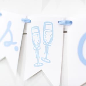 Op de afbeelding: Een witte wimpelslinger met lichtblauwe letters en een centrale afbeelding van twee champagneglazen. De afzonderlijke wimpels zijn verbonden door een lichtblauw lint dat door gaten aan de bovenkant van elk driehoekig papieren stuk is geregen.