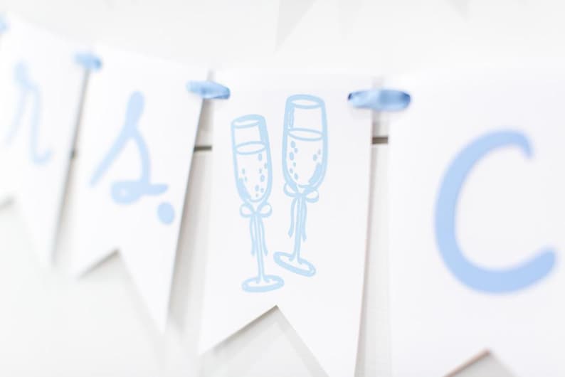 Op de afbeelding: Een witte wimpelslinger met lichtblauwe letters en een centrale afbeelding van twee champagneglazen. De afzonderlijke wimpels zijn verbonden door een lichtblauw lint dat door gaten aan de bovenkant van elk driehoekig papieren stuk is geregen.