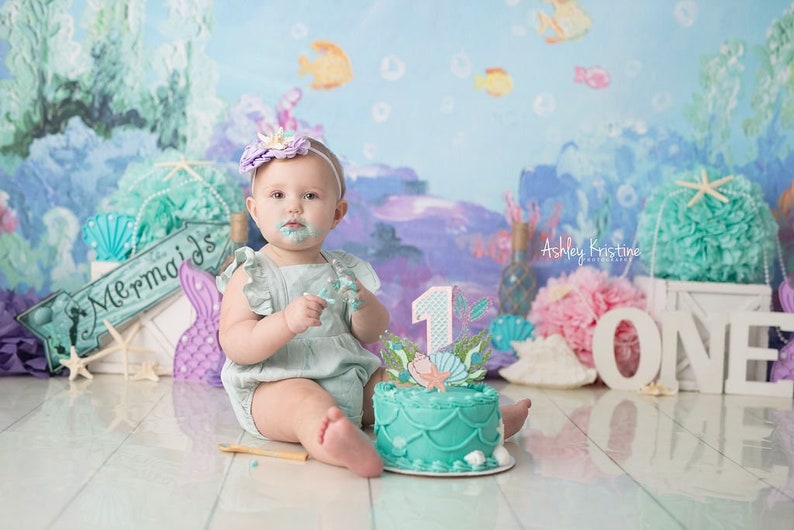 May include: A baby girl wearing a light blue dress and a purple flower headband sits in front of a mermaid-themed backdrop. She is holding a piece of cake and has frosting on her face. The cake is decorated with a mermaid tail and a number one. The backdrop features a sign that says "Mermaids" and other mermaid-themed decorations.