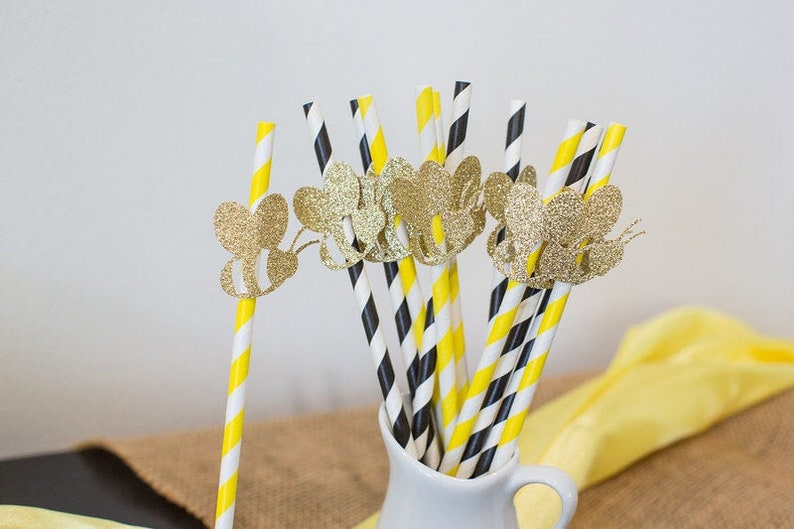 May include: A collection of decorative paper straws in a white pitcher. The straws feature yellow and white stripes, black and white stripes, and are adorned with gold glitter bee cutouts. A yellow cloth is in the background.