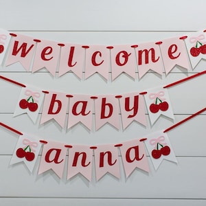 May include: A three-tiered banner with the words "welcome baby anna" in red script on pink pennants. Each pennant is adorned with a red cherry illustration. The banner is strung with red ribbon against a white wood-paneled background.