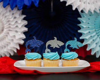 May include: Three cupcakes with blue frosting and glitter shark toppers on a white plate. The cupcakes are in front of a background of blue, white, and light blue paper fans.