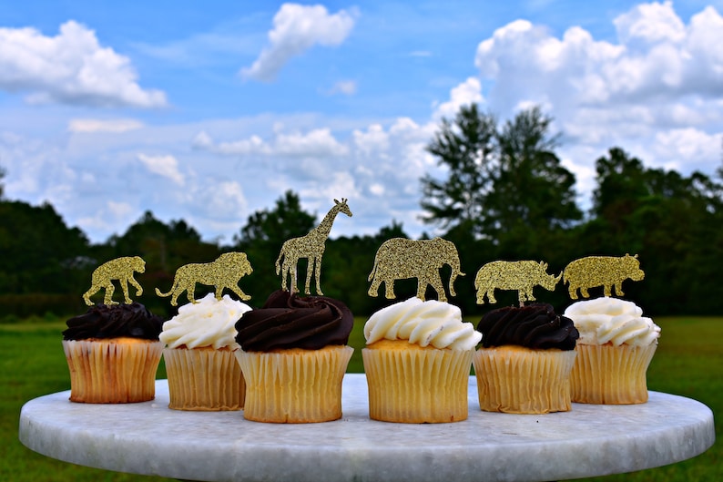 May include: Cupcakes with gold glitter animal toppers, including a lion, giraffe, and elephant. The cupcakes have vanilla and chocolate frosting. The treats are arranged on a white marble cake stand, with a green and blue sky background.