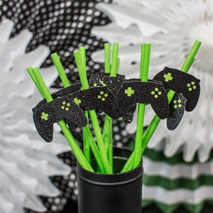 May include: A bunch of green straws with black and green glitter game controller cutouts. The straws are in a black vase against a background of white and black paper fans.
