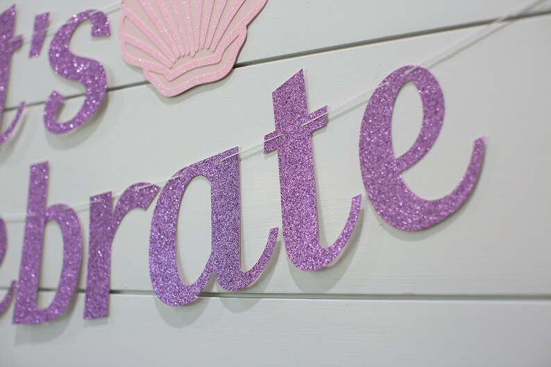 May include: Purple glitter banner with the words "Let's Celebrate" on a white wood background.