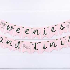 May include: Pink banner with the words "weenies and tinis" in dark green script. Each pennant is pink and has a small illustration of a dachshund and a martini glass. The banner is strung on green ribbon.