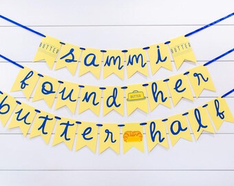 May include: A yellow banner with blue text that reads "Sammi found her butter half." The banner is strung with blue ribbon and has a white wooden background. The banner has a butter and croissant illustration.
