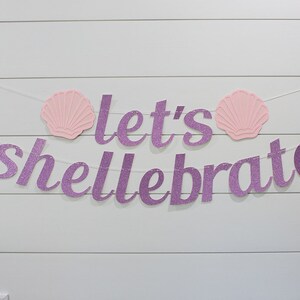 May include: Purple glitter banner with the text "Let's Shellebrate" and two pink seashells on a white wood background.