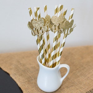 May include: A white ceramic pitcher filled with gold and white striped paper straws with gold glitter bee cutouts.