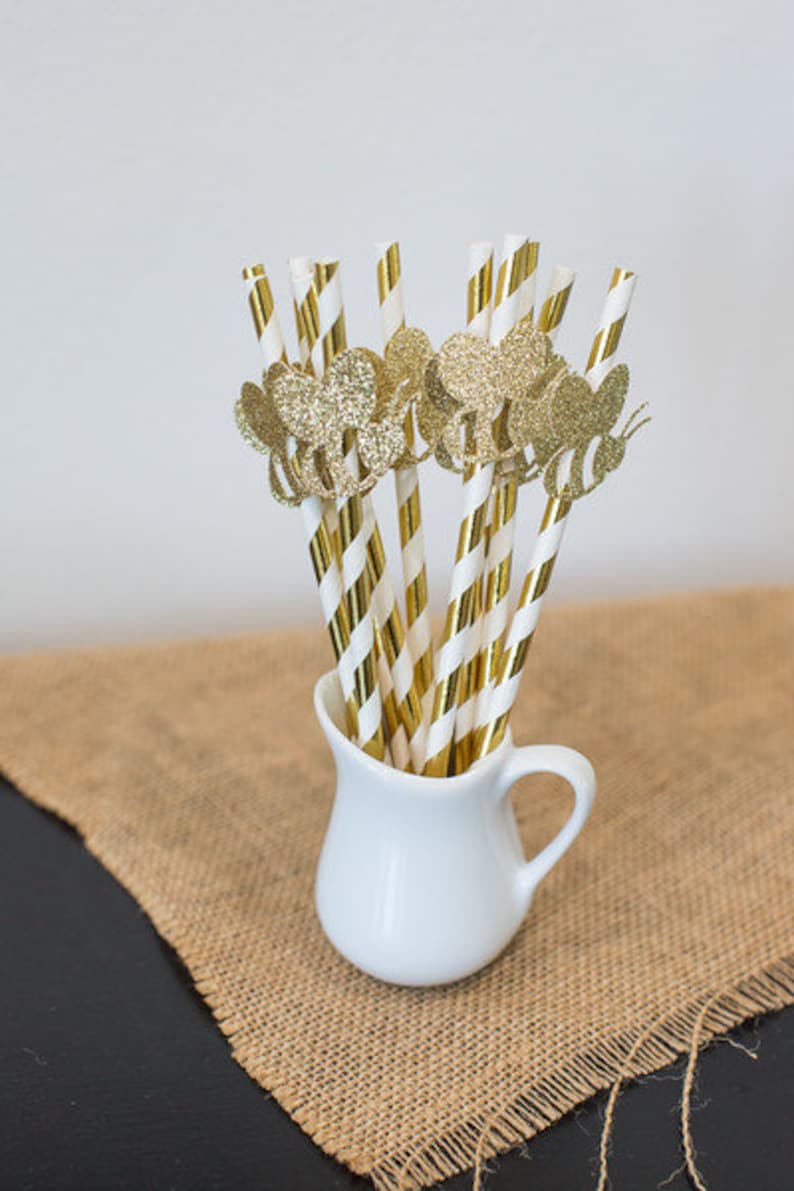 May include: A white ceramic pitcher filled with gold and white striped paper straws with gold glitter bee cutouts.