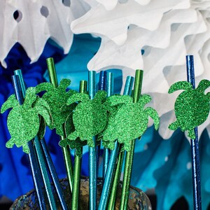 May include: A close-up of green glitter turtle straws. The straws are arranged in a bunch and are surrounded by blue and white paper decorations.