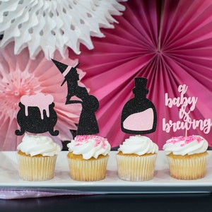 May include: Four cupcakes decorated with pink and black glitter. The cupcakes are topped with witch-themed toppers, including a pregnant witch, a cauldron, a potion bottle, and a sign that says "a baby is brewing".