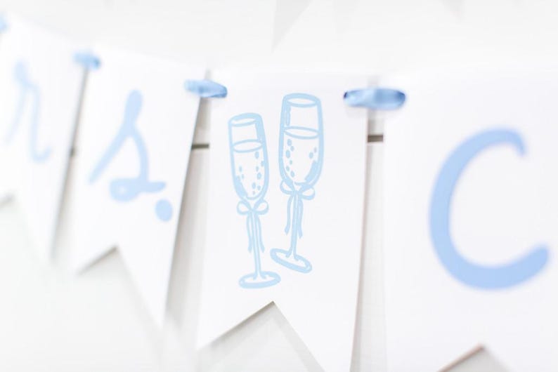 Op de afbeelding: Een witte wimpelslinger met lichtblauwe letters en een centrale afbeelding van twee champagneglazen. De afzonderlijke wimpels zijn verbonden door een lichtblauw lint dat door gaten aan de bovenkant van elk driehoekig papieren stuk is geregen.