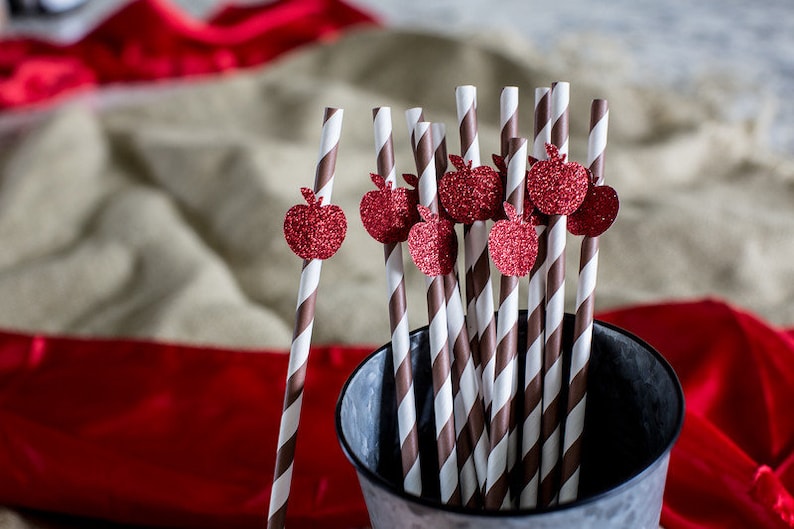 May include: A close-up of a bunch of brown and white striped paper straws with red glitter apple decorations on the top. The straws are in a silver metal bucket.