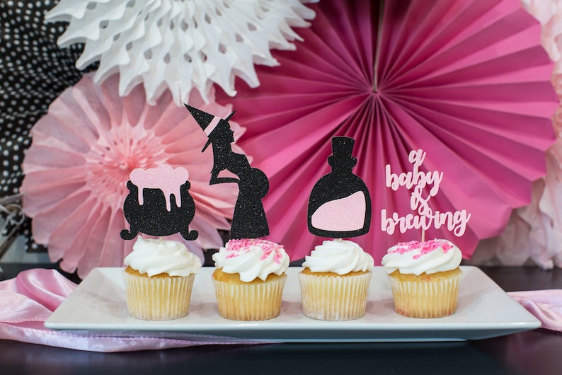 May include: Four cupcakes decorated with pink and black glitter. The cupcakes are topped with witch-themed toppers, including a pregnant witch, a cauldron, a potion bottle, and a sign that says "a baby is brewing".