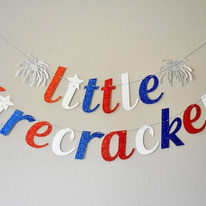 Pu&ograve; includere: Uno striscione di glitter rosso, bianco e blu che recita "little firecracker". Lo striscione &egrave; infilato su un filo bianco e ha forme di fuochi d'artificio di glitter argentati a ciascuna estremit&agrave;.