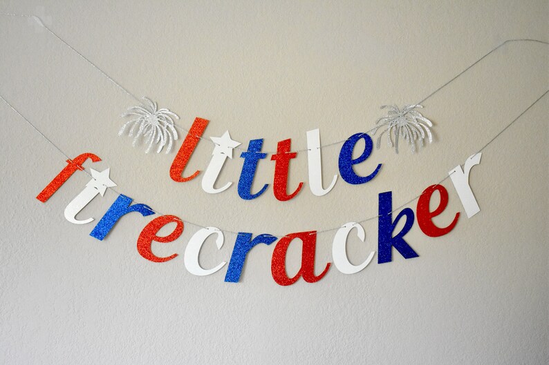 Pu&ograve; includere: Uno striscione di glitter rosso, bianco e blu che recita "little firecracker". Lo striscione &egrave; infilato su un filo bianco e ha forme di fuochi d'artificio di glitter argentati a ciascuna estremit&agrave;.