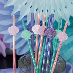 May include: A bunch of colorful straws with glitter seashells attached to the top. The straws are in a clear glass jar. The background is blue and pink.