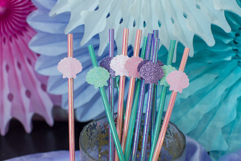 May include: A bunch of colorful straws with glitter seashells attached to the top. The straws are in a clear glass jar. The background is blue and pink.