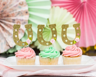 May include: Three cupcakes decorated with gold glitter horseshoe toppers. The cupcakes are frosted in pink and green.