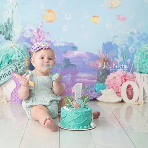May include: A baby girl wearing a light blue dress and a purple headband with a seashell design is sitting on a white wooden floor. She is holding a wooden spoon and eating cake. The cake is decorated with a mermaid and the number one. The background is a blue and purple ocean-themed backdrop with the words "Mermaids" and "ONE" in white letters.