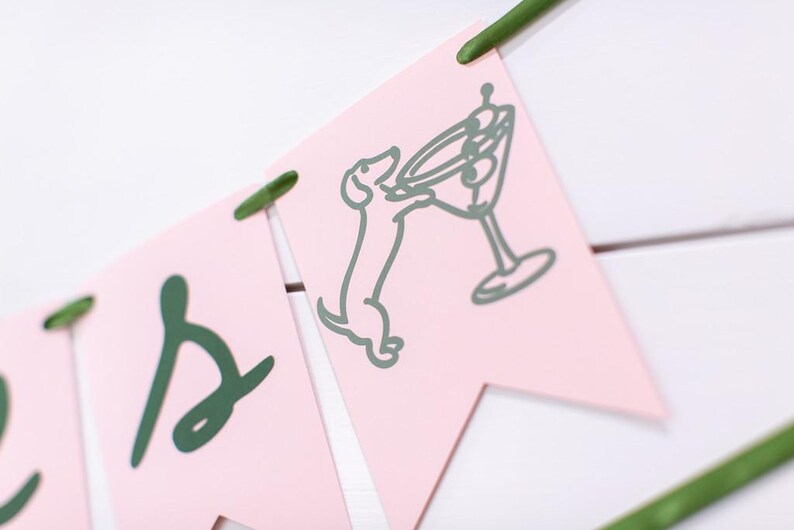 May include: Pink banner with green lettering and illustrations. One banner features a dachshund dog reaching for a martini glass. The banner is strung together with green string and is set against a white background.