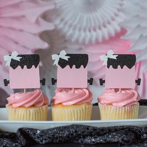 May include: Three pink cupcakes with black and white frosting toppers. The toppers are shaped like a square with a black top and a pink bottom. Each topper has a white bow on top.