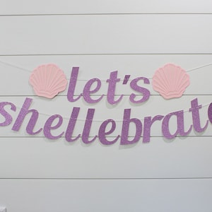May include: Purple glitter banner with the text "Let's Shellebrate" and two pink seashells on a white wood background.