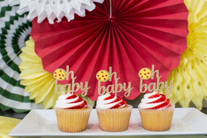 May include: Three yellow and white cupcakes with red frosting and gold glitter "Oh Baby" toppers featuring a yellow and red polka dot design.