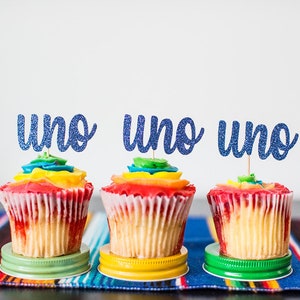 May include: Three cupcakes with rainbow frosting and blue glitter "uno" toppers. The cupcakes sit on colorful jar lids atop a striped blanket. The cupcakes have red, yellow, and green frosting.