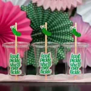 May include: Three clear plastic cups with the text "last pickle she'll ever tickle" in green. Each cup has a wooden stick with a green glitter pickle and white bow. Pink and green paper fans are in the background.