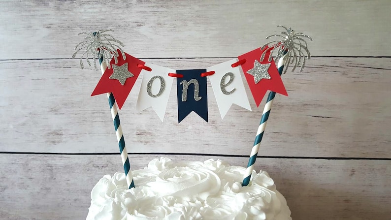 Pu&ograve; includere: Un topper per torta con uno striscione rosso, bianco e blu che dice "one" con stelle argentate e disegni di fuochi d'artificio. Lo striscione &egrave; attaccato a due cannucce a righe.