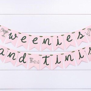 May include: Pink banner with the words "weenies and tinis" in dark green script. Each pennant is pink and has a small illustration of a dachshund dog and a martini glass. The banner is strung on green ribbon.