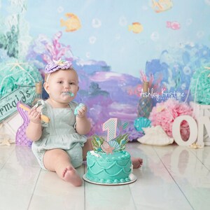 May include: A baby girl wearing a light blue dress and a purple headband with a seashell design is sitting on a white wooden floor. She is holding a wooden spoon and eating cake. The cake is decorated with a mermaid and the number one. The background is a blue and purple ocean-themed backdrop with the words "Mermaids" and "ONE" in white letters.