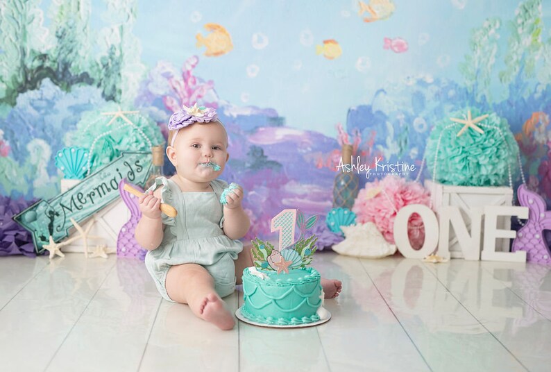 May include: A baby girl wearing a light blue dress and a purple headband with a seashell design is sitting on a white wooden floor. She is holding a wooden spoon and eating cake. The cake is decorated with a mermaid and the number one. The background is a blue and purple ocean-themed backdrop with the words "Mermaids" and "ONE" in white letters.