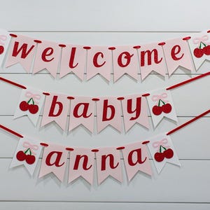 May include: A three-tiered banner with the words "welcome baby anna" in red script on pink pennants. Each pennant is adorned with a red cherry illustration. The banner is strung with red ribbon against a white wood-paneled background.