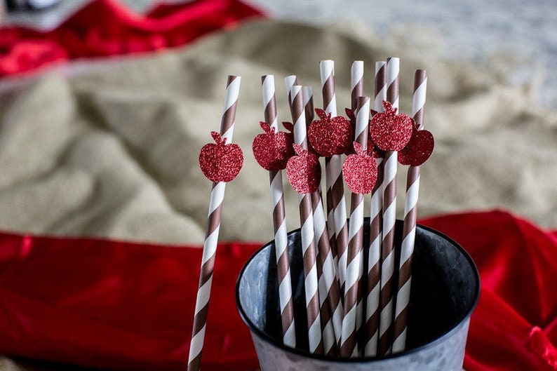 May include: A close-up of a bunch of brown and white striped paper straws with red glitter apple cutouts attached to the top. The straws are in a silver metal bucket.