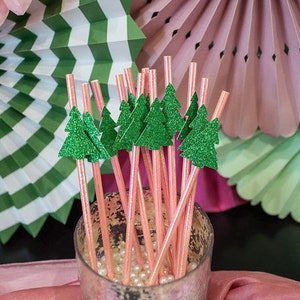 May include: A close-up of a glass jar filled with pink and white striped paper straws with green glitter Christmas tree toppers. The jar is sitting on a pink and white tablecloth.